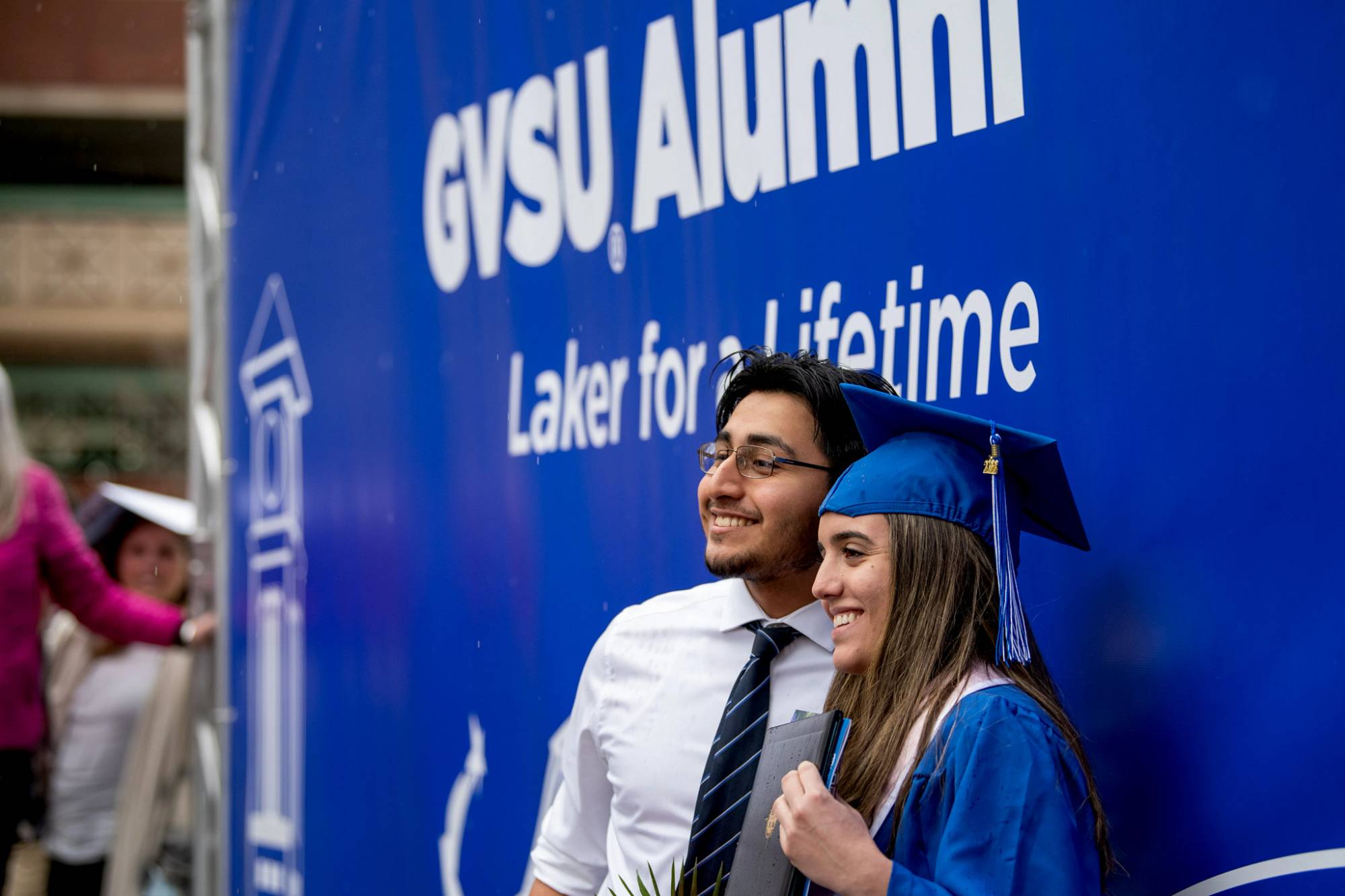 A grad stands in front of the GVSU Alumni banner with a family member
